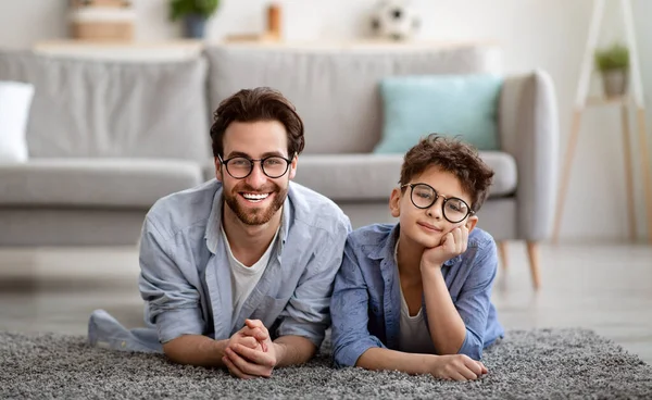 Portret van vrolijke vader en zoon met een bril op en lachend naar de camera terwijl ze op vloertapijt liggen — Stockfoto