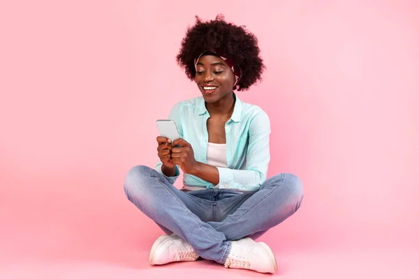 Αφρικανική γυναίκα που χρησιμοποιεί το τηλέφωνο κάθεται στο πάτωμα, ροζ φόντο — Φωτογραφία Αρχείου