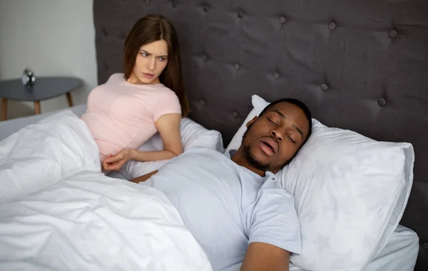 Ένας νεαρός μαύρος ροχαλίζει και ενοχλεί τη θυμωμένη άγρυπνη γυναίκα του στο κρεβάτι στο σπίτι. — Φωτογραφία Αρχείου
