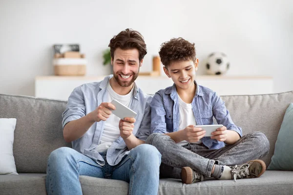 Ontspanning en vrije tijd. Vader concurreren met zoon in online video games op smartphones, samen zitten op de bank — Stockfoto