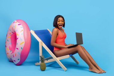 Yaz indirimi. Mayo giymiş heyecanlı bir zenci kadın dizüstü bilgisayarla şezlongda oturmuş mavi arka planda OMG diye bağırıyor.