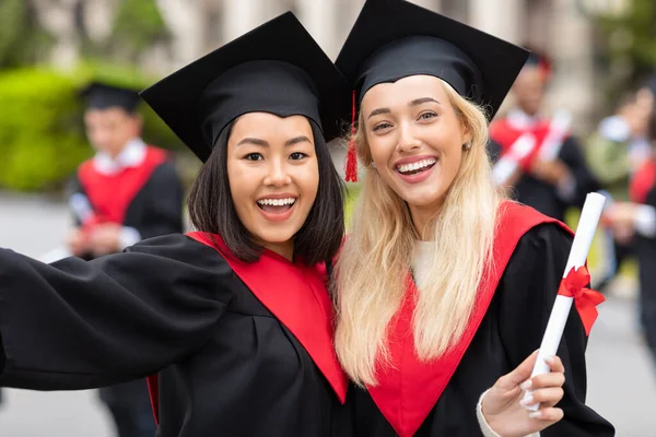 Jovens senhoras multirraciais felizes em roupas de graduação levando selfie — Fotografia de Stock