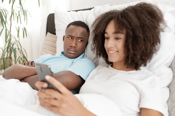 Young couple having problem. Jealous husband is watching his wife use mobile phone on bed at home — Stock Photo, Image