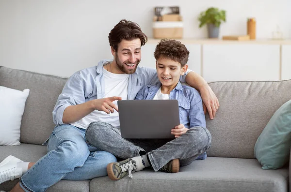 Papa attentionné enseignant fils à utiliser un ordinateur portable ou naviguer sur Internet ensemble, assis sur un canapé confortable dans le salon — Photo