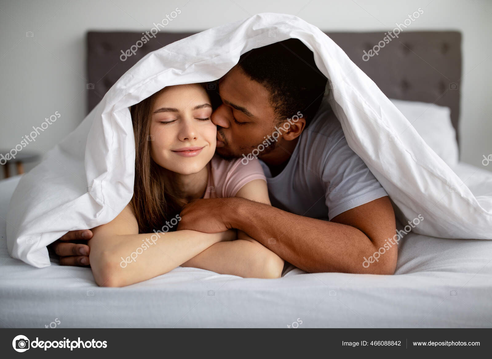 Romantic black guy kissing his Caucasian girlfriend under blanket on bed, indoors Stock Photo by ©Milkos 466088842