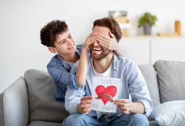 Babalar Günü 'nde babasına sürpriz yapan mutlu bir oğul. Gözlerini arkadan kapatıyor, selamlıyor ve el yapımı tebrik kartı veriyor.