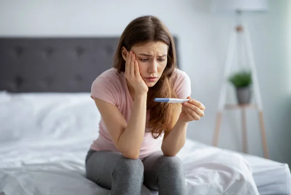 Traurige junge Frau überprüft ihren jüngsten Schwangerschaftstest und sitzt zu Hause im Bett — Stockfoto
