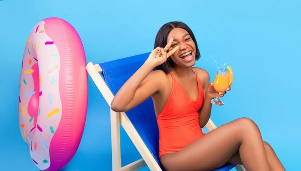 Yaz serinliğinde şezlong. Güzel siyah kadın tropikal kokteyl içiyor ve mavi arka planda barış jesti yapıyor. — Stok fotoğraf