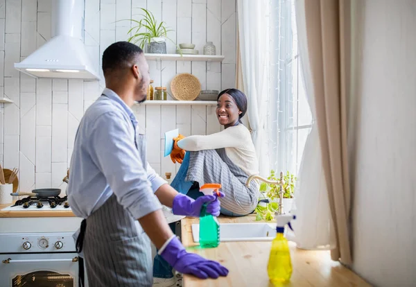 Jeune couple prenant une pause pendant le nettoyage de la maison, homme noir debout avec du détergent, petite amie assise sur la table de cuisine — Photo