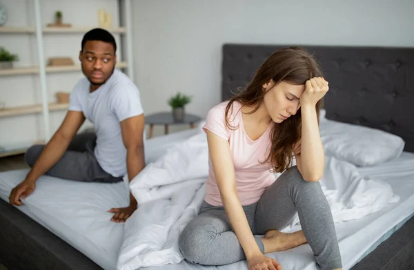 Multiracial couple having relationship difficulties, sitting on bed, feeling upset, going through marital crisis at home