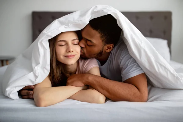 Romantico ragazzo nero che bacia la sua ragazza caucasica sotto la coperta sul letto, al chiuso — Foto Stock