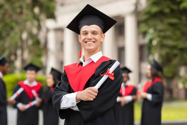 Веселий хлопець у випускному костюмі з дипломом позує на відкритому повітрі — стокове фото
