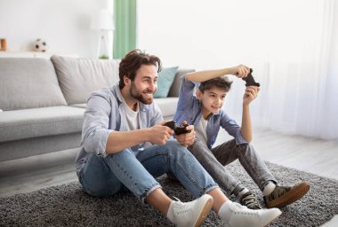 Video oyunları konsepti. Neşeli baba ve oğul çevrimiçi oyunlarda yarışıyor, halının üzerinde otururken joystickle oynuyorlar.