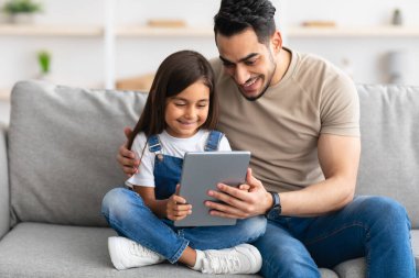 Gülümseyen baba ve kız kanepede oturuyor, tablet kullanıyor.