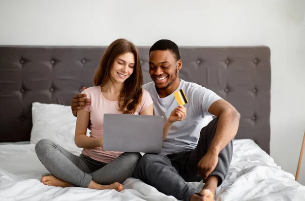 Positivt rasblandat par med kreditkort och laptop shopping online på sängen hemma — Stockfoto
