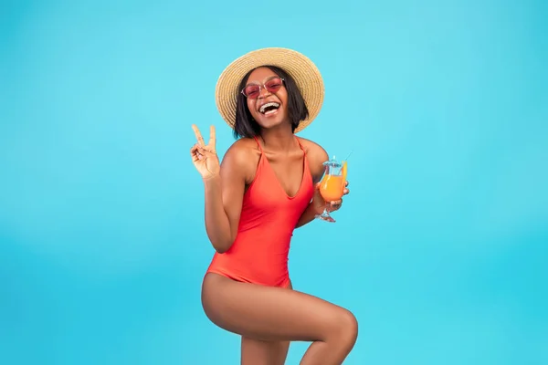 Mulher negra feliz em maiô desfrutando de coquetel de verão exótico e mostrando sinal de paz, posando sobre fundo azul — Fotografia de Stock