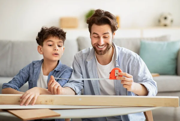 Молодий тато і його підліток син вимірюють дерев'яну дошку зі стрічковим лінійкою, чоловік навчає хлопчика робити домашнє завдання — стокове фото