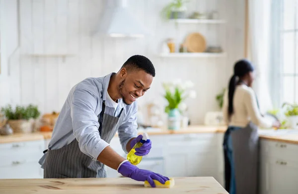 Bello ragazzo nero in guanti di gomma pulizia tavolo da cucina con detergente, sua moglie lavare i piatti su sfondo — Foto Stock