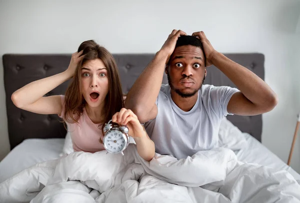 Casal multirracial chocado com despertador sendo tarde para o trabalho, sentado na cama, agarrando suas cabeças em terror — Fotografia de Stock