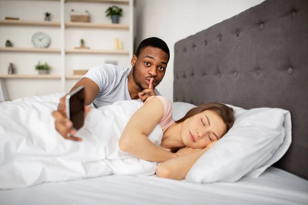 Féltékeny fekete férj kémkedik a felesége után, HUSH gesztust mutat, ellenőrzi a mobilját, miközben alszik az ágyban. — Stock Fotó