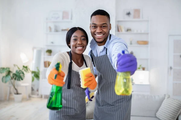 Cara preto feliz e sua esposa apontando detergentes spray para a câmera, realizando limpeza doméstica, dentro de casa — Fotografia de Stock