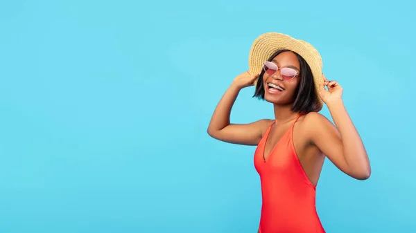 Happy black lady in bikini wearing sunglasses and straw hat, posing on blue background, banner design with free space — Stock Photo, Image