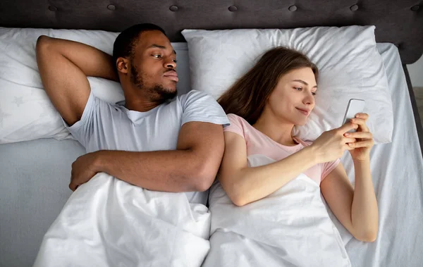 Irritierter Schwarzer, der eifersüchtig auf seine Freundin ist, die im Bett im Smartphone steckt, von oben gesehen. Phubbing-Konzept — Stockfoto