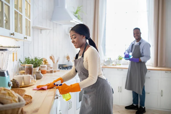 Mulher negra alegre limpeza mesa de cozinha, seu namorado lavar pratos no fundo, espaço em branco — Fotografia de Stock