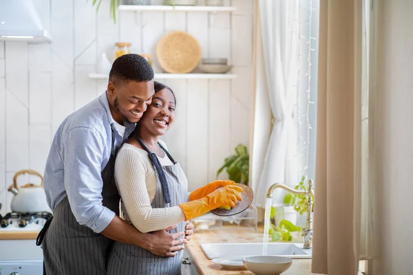 Hombre negro guapo abrazando a su novia mientras ella está lavando platos en casa, espacio libre — Foto de Stock
