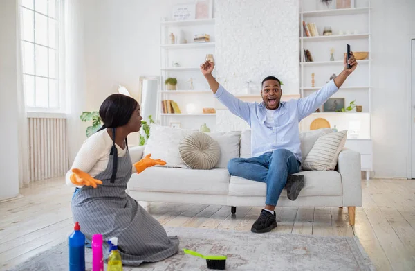 Ενθουσιασμένος μαύρος που βλέπει ποδόσφαιρο στην τηλεόραση, χαρούμενος για τη νίκη της ομάδας του, θυμωμένος προσβεβλημένος σύζυγος καθαρίζει το σπίτι μόνος του. — Φωτογραφία Αρχείου