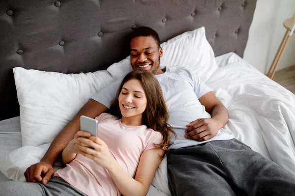 Feliz casal interracial deitado na cama com smartphone, navegando na web, tendo videochamada on-line, verificando as mídias sociais — Fotografia de Stock