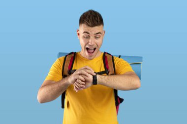 Endişeli genç adam kamp malzemeleriyle saatine bakıyor, mavi stüdyo geçmişini kontrol ediyor.