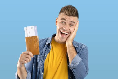 Uluslar arası pasaportu ve mavi stüdyo arka planında uçak biletleri olan şoke olmuş bir adam.
