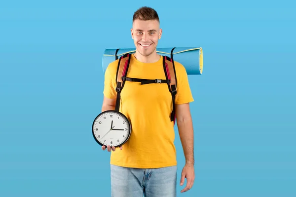 Czas na koncepcję podróży. Przystojny millenium facet z plecakiem trzyma zegar nad niebieskim tle studio — Zdjęcie stockowe