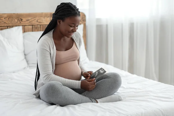Щаслива чорна вагітна жінка з ультразвуковою фотографією дитини, що сидить на ліжку — стокове фото