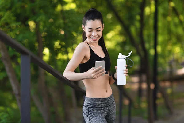 Menina asiática nova usando Smartphone e água potável, descansando após o exercício ao ar livre — Fotografia de Stock