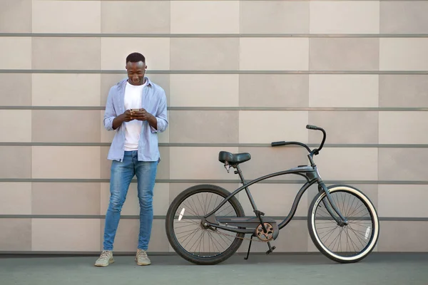 Šťastný černý biker ve sluchátkách procházení internetu pro novou hudbu na mobilu v blízkosti cihlové zdi, kopírovat prostor — Stock fotografie