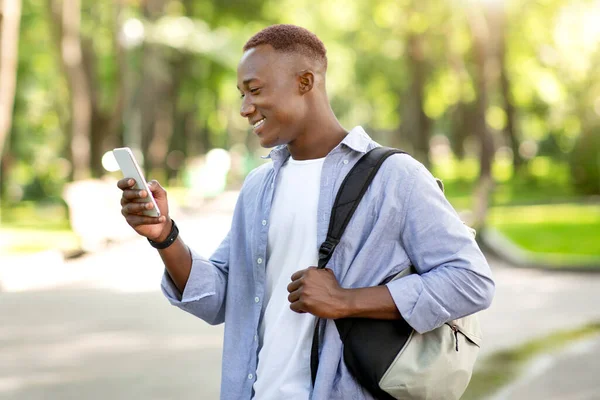 Alegre hombre negro con mochila usando el teléfono celular a pie en el parque de la ciudad — Foto de Stock