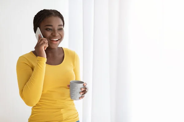 Excited Black Woman With Cellphone And Coffee Near Window Enjoying Phone Call