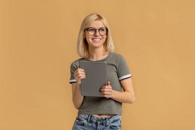Elinde dijital tabletle mutlu bir kadın gülümsüyor, hayal kuruyor ve uzaklara bakıyor, bej arka planda poz veriyor.