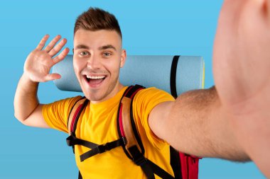 Kamp vitesleriyle milenyum adamı selfie çekiyor ve mavi stüdyo arka planında akıllı telefon kamerasına el sallıyor.