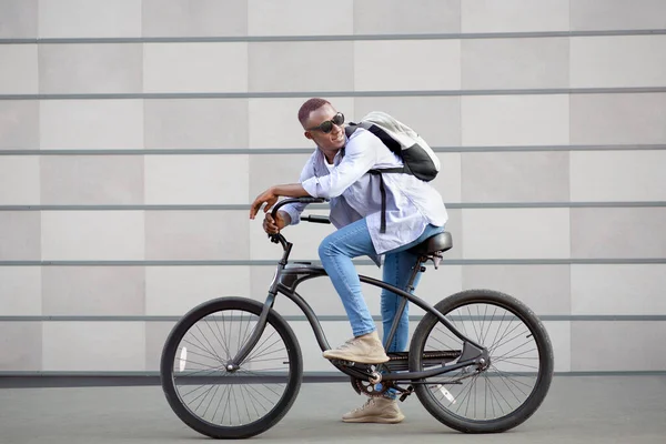 Retrato de cara preto bonito com mochila e óculos de sol andando de bicicleta perto da parede de tijolo fora — Fotografia de Stock