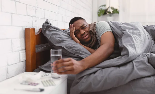 Africano cara tomando medicação tendo febre deitado na cama interior — Fotografia de Stock