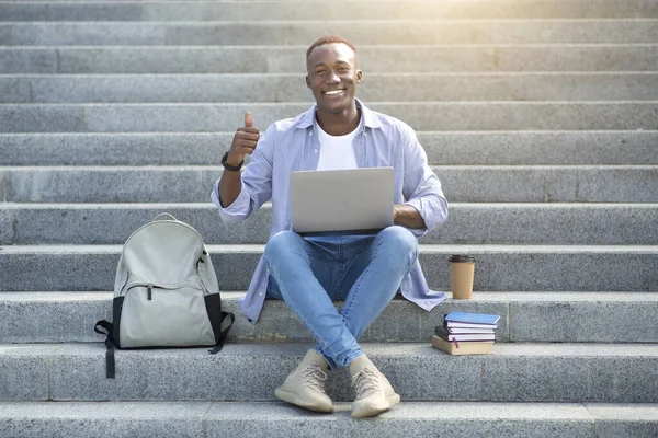 Positive black man with laptop showing thumb up gesture on stairs at city centre