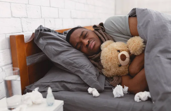 Λυπημένος άρρωστος Αφρικανός που αγκαλιάζει αρκουδάκι ξαπλωμένο στην κρεβατοκάμαρα. — Φωτογραφία Αρχείου