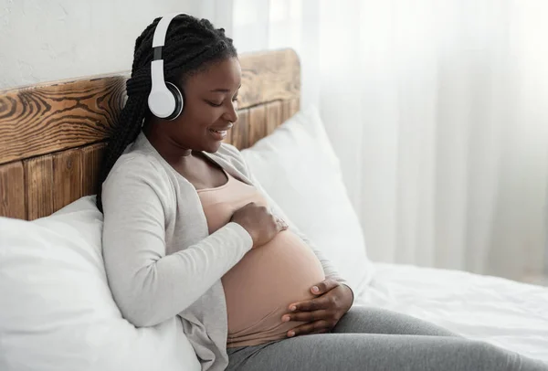 Šťastná černošky těhotná žena ležící v posteli, poslech hudby v bezdrátových sluchátkách — Stock fotografie