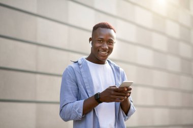 Kulaklıklı Afro-Amerikalı adam şehir merkezindeki tuğla duvarın yanında cep telefonundan müzik dinliyor.