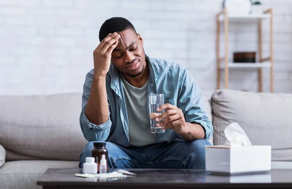 Несчастный черный парень страдает от головной боли, принимая таблетки, сидя в помещении — стоковое фото