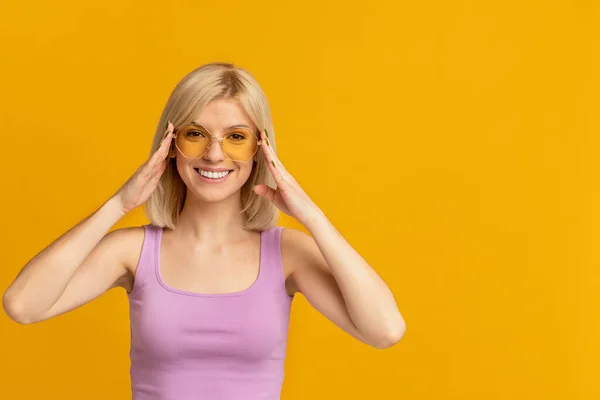 Mujer atractiva positiva sonriendo y usando gafas de sol con estilo, posando sobre fondo de estudio amarillo —  Fotos de Stock