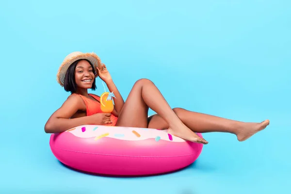 Sexy dama negra sentada en el anillo de rosquilla inflable, bebiendo cóctel de verano sobre fondo azul, retrato de cuerpo entero — Foto de Stock